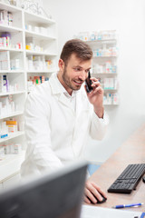 Smiling handsome young male pharmacist talking on the phone and working on a computer. Standing behind the counter in pharmacy. Healthcare and medicine concept.