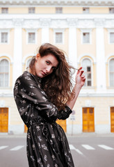 young beautiful woman in a dress with wavy hair stands on the street against the background of a building, portrait,