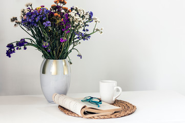 bouquet of flowers, open book and glasses, lemon tea in a white ceramic cup on a wicker stand on the table, cozy home interior.