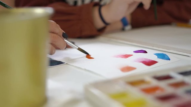 Girl draws on white brown with brush, violet paint drawing on Mester class in fine arts. Multi-colored watercolor palette colored paints. Many people. Learn to draw