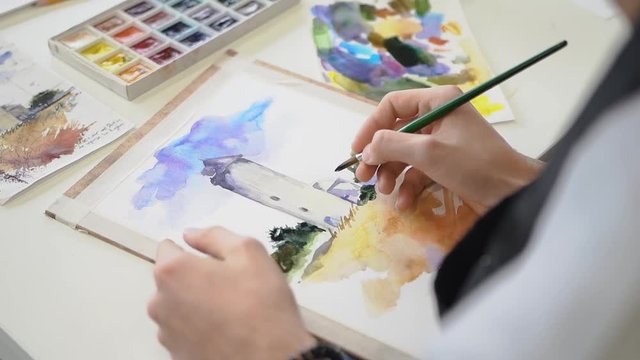 The girl draws on white paper with a brush and blue paint a drawing on the Mester class in the fine arts. Multi-colored watercolor palette with colored paints.