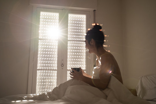 A beautiful dark-haired woman is in her bed while take a cup of coffee. She is smiling. The sunlight go into the room through the window. She is cover with a eiderdown.