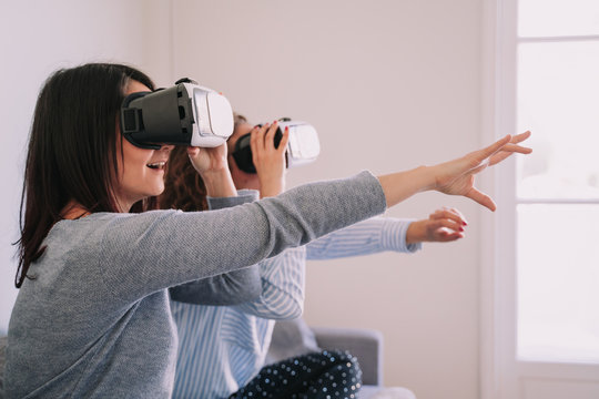 Two beautiful girls are sitting in the armchair at home playing with virtual reality glasses. They look through them and try to reach for something in front of them by extending their arms.