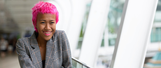 Happy young rebellious Asian businesswoman with pink hair looking at view of the city
