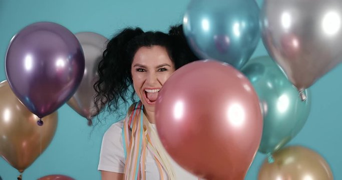 Portrait of beautiful young woman with black curly hair smiling and dancing with ballons in her hands in front of blue background shot in 4k super slow motion