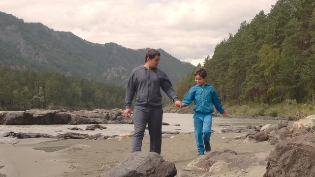 Father And His Young Son Are Walkng Along The Riverside In Mountains In Summer