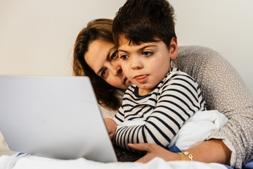 una mujer madre y su hijo con pluridiscapacidad (minusvalía, discapacidad) juegan con el ordenador portátil o tablet en la cama. van en pijama, y están alegres y sonrientes.