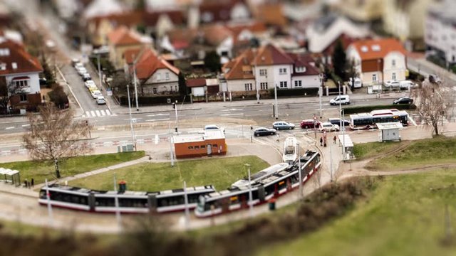 Aerial view of the tram stop and a crossroad near Divoka Sarka park in Prague. Active traffic on the road, cars, busses, and trams. Commuters crowd the tram stop. Toylike houses with orange roofs.