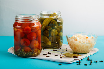 Jars with pickled cucumbers and tomatoes and sauerkraut on a wooden table on a blue background on a linen towel.