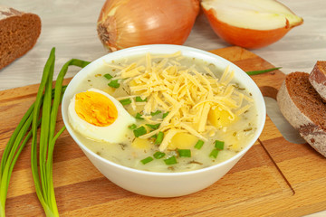 A white plate of cheese and potato onion soup with grated cheese poured on top and half a boiled egg surrounded by slices of rye bread, green onions and onions.