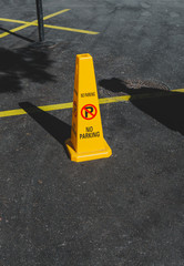 Yellow road cone with a warning not to park on a back of gray asphalt road.