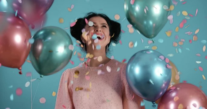 Portrait of beautiful young woman with black curly hair smiling dancing with ballons in her hands raining confetti in front of blue background shot in 4k super slow motion