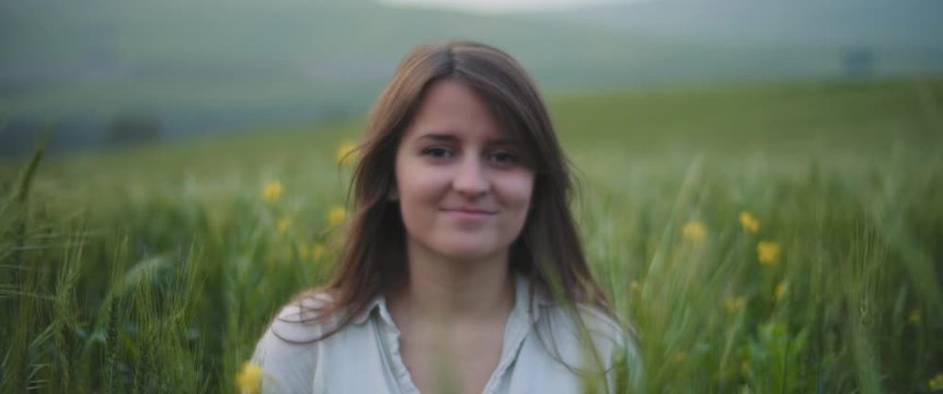 A young woman with long brown hair in a middle of a green wheat field, smiling, feeling happy. CLOSE UP, SLOW MOTION, SHALLOW DOF. Freedom, lifestyle concept. BMPCC 4K 