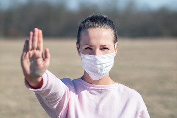 Girl, young woman in protective sterile medical mask on her face looking at camera outdoors, show palm, hand, stop no sign. Air pollution, virus, Chinese pandemic coronavirus concept. Covid-19