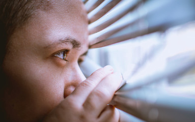 man warily looks out the window into the street through the blinds while staying at home during the...