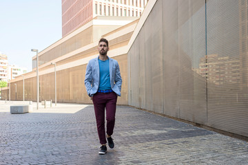 Fototapeta na wymiar Young bearded man, model of fashion, in urban background wearing casual clothes while walking with hands on pocket