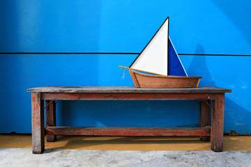 Small wooden ship toy model on long wooden chair in blue concrete background, summer vacation concept 