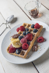 Homemade waffle with banana slices, oat, pecans and berries served on a white plate over a white rustic wooden table. Closeup.