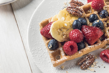 Homemade waffle topped with banana slices, oat, pecans and berries served on a white plate over a white rustic wooden table. Closeup with copyspace.