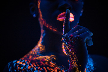 ethnic fluorescence patterns on cropped young woman's body, skin. creative body art, in neon lights. unusual shoot of woman showing silence gesture