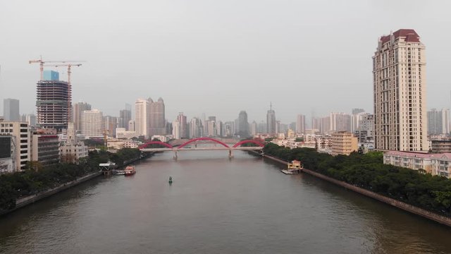 Green banks of Pearl river, red tied-arch Jiefang bridge seen ahead, aerial shot of Guangzhou. Center of city built with tall and low-rise buildings. Two piers on opposite banks for short ferry route