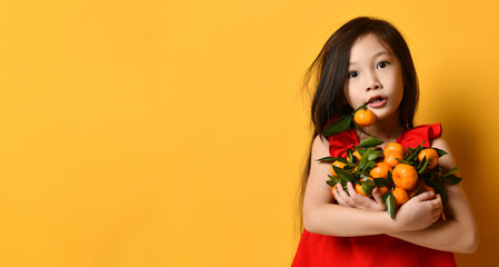 Asian girl in red blouse. Looking surprised, holding an armful of tangerines and oranges, posing on orange background. Close up