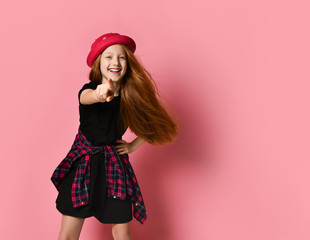 Red-haired teenage child in black dress, checkered shirt on waist, red hat. Pointing at you, posing on pink background. Close up