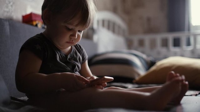 Focused European chubby baby boy watching video on smartphone, playing with phone while sitting on couch in cozy living room. Concept of modern entertainment young children. Shooting in slow motion.