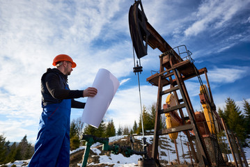 Petroleum engineer in blue uniform and orange helmet standing in oil field with printed plan next to the oil pump jack, on beautiful sunny day. Concept of petroleum industry and oil extraction.