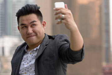 Young overweight Asian businessman taking selfie against view of the city