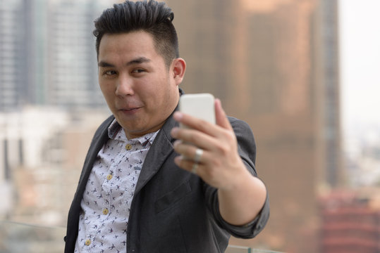Young overweight Asian businessman taking selfie against view of the city