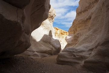 The narrow canyon with high white sandstone walls, big stones and dark shadow on its bottom. There is the blue sky with white clouds.