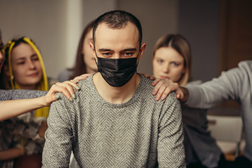 ill caucasian guy in medical protective mask suffer from incurable virus infection, sit in center of isolated room with strangers supporting him in such hard time. sickness, cancer, coronavirus