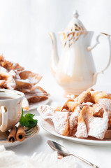 Chenchi carnival sweets on a white plate. Sprinkled with powdered sugar. Nearby is a cup of tea decorated with cinnamon and mint. In the background is a porcelain teapot. White background. 