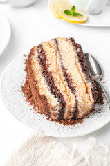 A piece of sponge cake with a chocolate layer soaked in butter cream. Home made. On a white plate. Nearby is a white porcelain cup. Close-up. On a white linen tablecloth.