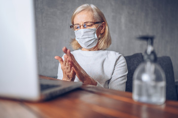 Lonely sad older senior woman in face medical mask using hand antibacterial liquid sanitizer with laptop computer at home self isolation quarantine during coronavirus COVID19 pandemic. Stay home 