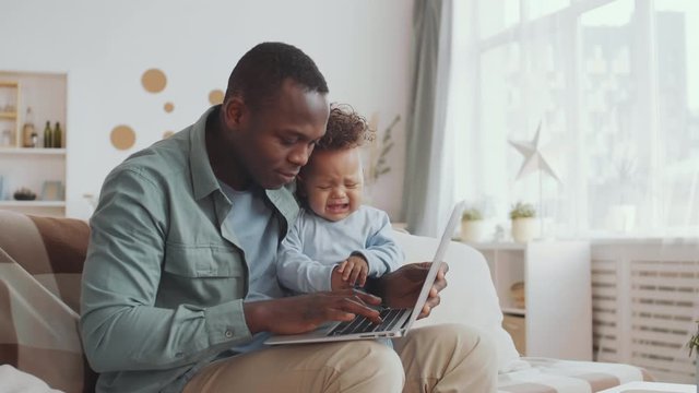 Medium shot of handsome African man sitting on couch in living room, holding active curly-haired kid while networking using laptop