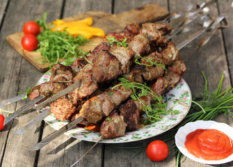 Grilled pork shashlik on skewers on a light plate on an old wooden background with ketchup in a bowl, tomatoes, peppers and herbs. Background image, copy space