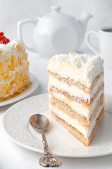 A piece of sponge cake with whipped cream and berries. garnished with pieces of white chocolate. Home made. On a white plate. In the background is a cake, a teapot and a white mug. 