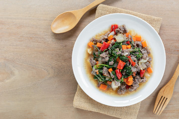 top view of stir fried minced beef with holy basil in a ceramic dish on wooden table. hot and spicy thai style food menu.