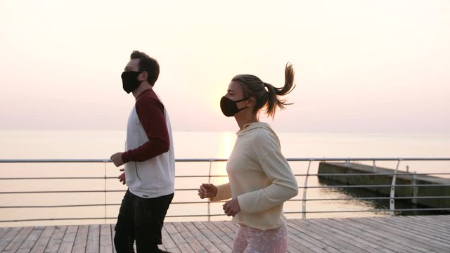 Young fit couple in protective masks running outdoors near the sea during beautiful sunrise, slow motion