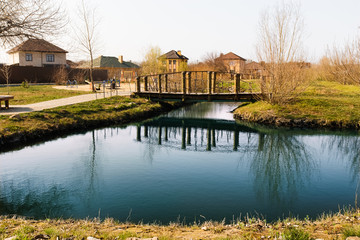 Obraz premium Blue pond with sky reflection and village houses.Wooden bridge over a small river or pond, side view. Pedestrian bridge over the river on
