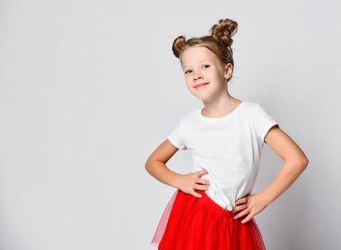 Cheerful kid girl in red fatin skirt and white t-shirt is standing holding hands on her waist to us having fun dancing