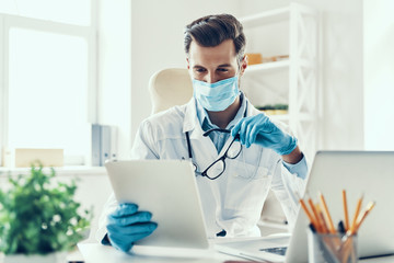 Busy young man in white lab coat and protective mask working usi