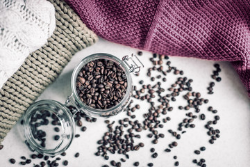Macro shot of a transparent jar filled with coffee beans in the center of the photo, coffee beans around the jar and knitted sweaters in the background 