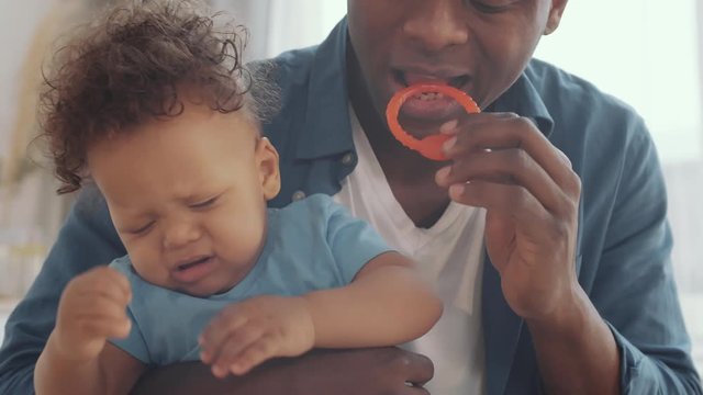 Close up of attractive African man sitting and holding crying baby boy on his hands and trying to calm him down using different toys