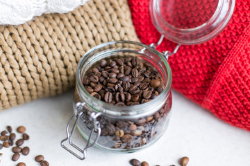 Macro shot of a transparent jar filled with coffee beans in the center of the photo, coffee beans around the jar and knitted sweaters in the background 