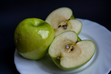 One whole green sour apple and two halves of the apple on white plate on dark background