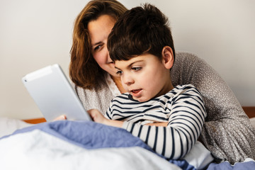 una mujer madre y su hijo con pluridiscapacidad (minusvalía, discapacidad) juegan con el ordenador portátil o tablet en la cama. van en pijama, y están alegres y sonrientes.