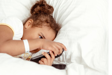Obraz na płótnie Canvas Cute little kid girl watching video on smartphone with smiley face alone on the bed, child using mobile phone with happy face at home. Stay at home quarantine coronavirus COVID-19 pandemic prevention.